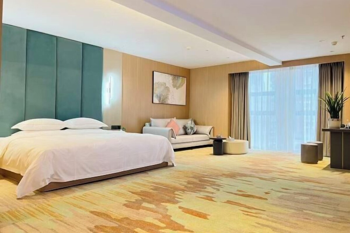 Una habitación de hotel con una cama grande, un sofá, una mesita y una ventana con cortinas.