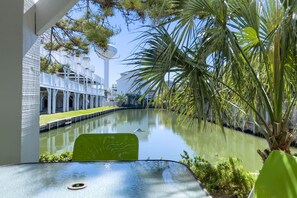 A view of a canal with a green chair in the foreground, a white building with balconies in the background, and a palm tree on the right.