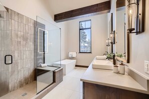 A modern bathroom with a glass shower enclosure, a bathtub, a vanity with a mirror, and a window providing natural light.