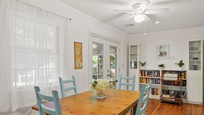 A dining area with a wooden table, blue chairs, a ceiling fan, and a bookshelf filled with books. - Artist's Cottage - Comfortable Pet friendly home 1 mile from the beach! (St Simons Island)