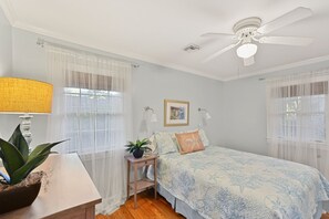 Une chambre à coucher avec un lit, une table de chevet, une lampe, un ventilateur de plafond et des rideaux légers.