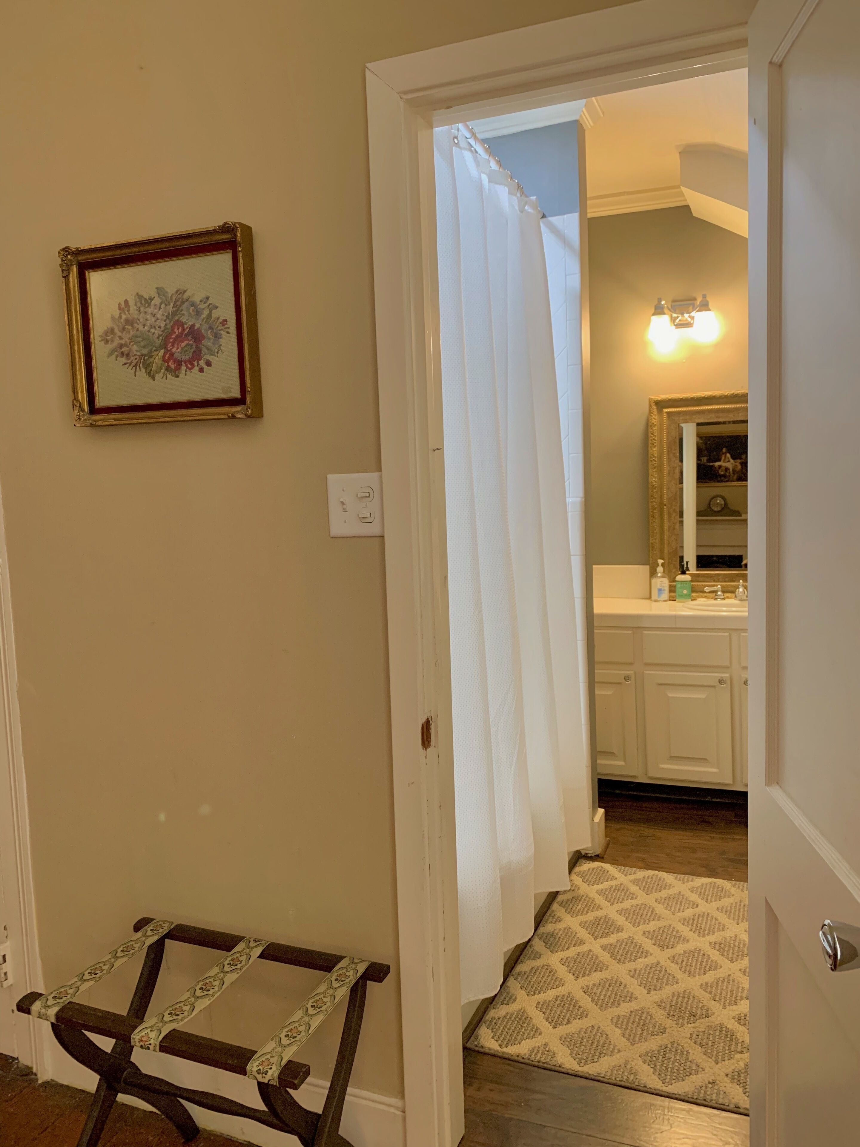 A hallway leading to a bathroom with a shower curtain, a framed floral artwork, and a patterned rug.