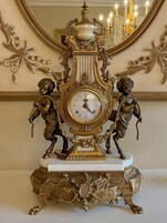 Un reloj de pie ornamentado con dos querubines, frente a una pared con molduras decorativas y un espejo.