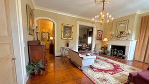 A spacious living room with hardwood floors, a chandelier, a fireplace, and a dining area in the background. - The Beaumont House (Natchez)
