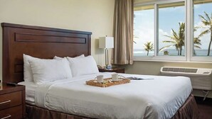 Habitació d'hotel amb un llit gran, una taula de nit amb una làmpada, una bandeja amb tasses i un llibre, i vista a palmeres per la finestra.