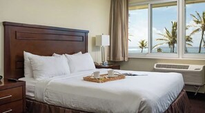 A hotel room with a large bed, a bedside table with a lamp, a tray with cups and a book, and a view of palm trees through the window.