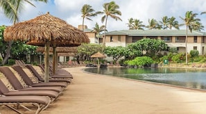 A resort pool area with lounge chairs and thatched umbrellas, surrounded by palm trees and a building with a green roof.