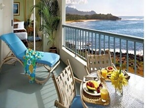 A balcony with a view of the ocean, a dining table with fruit and juice, and a lounge chair.