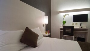 Una habitación de hotel con una cama, un escritorio con computadora, una silla y una mesita con lámpara y planta.