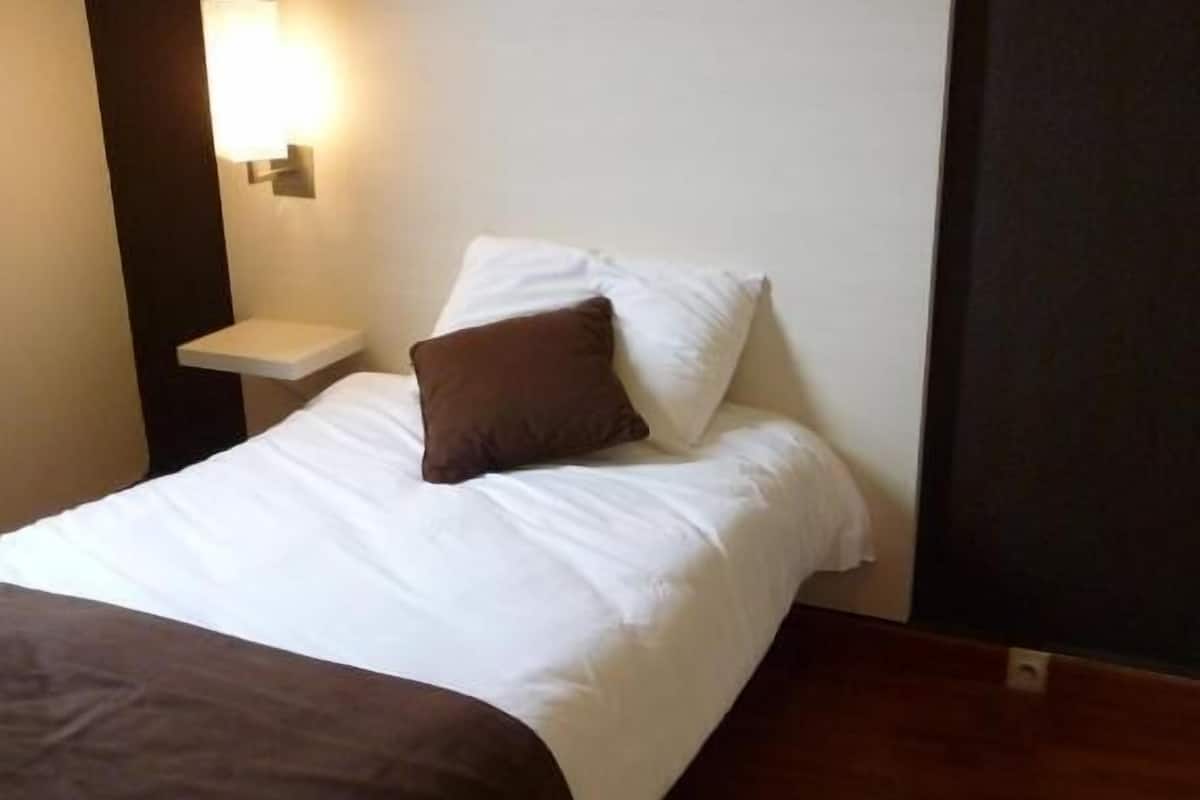 Una cama bien tendida con ropa blanca y un cojín marrón, una mesita de noche con una lámpara y un suelo de madera oscura.