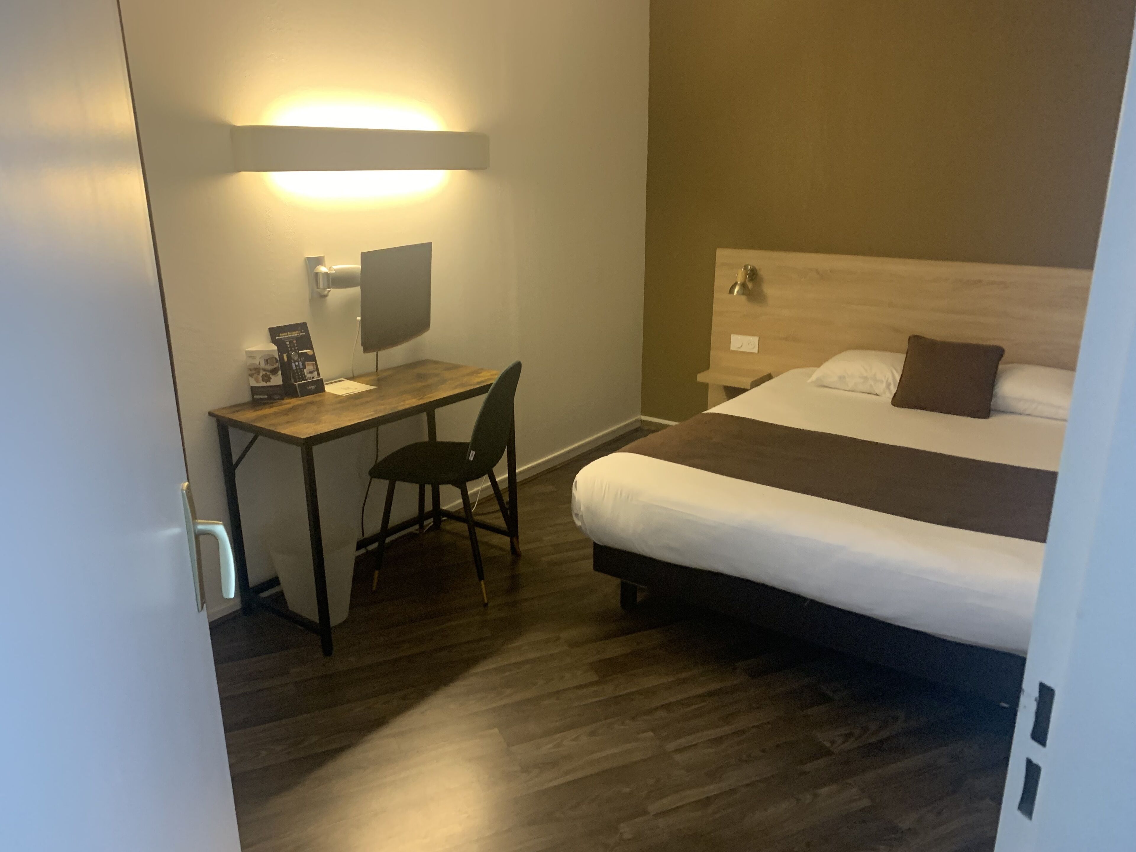 Quarto de hotel com escrivaninha de madeira, uma cadeira, cama com cabeceira e mesa de cabeceira.