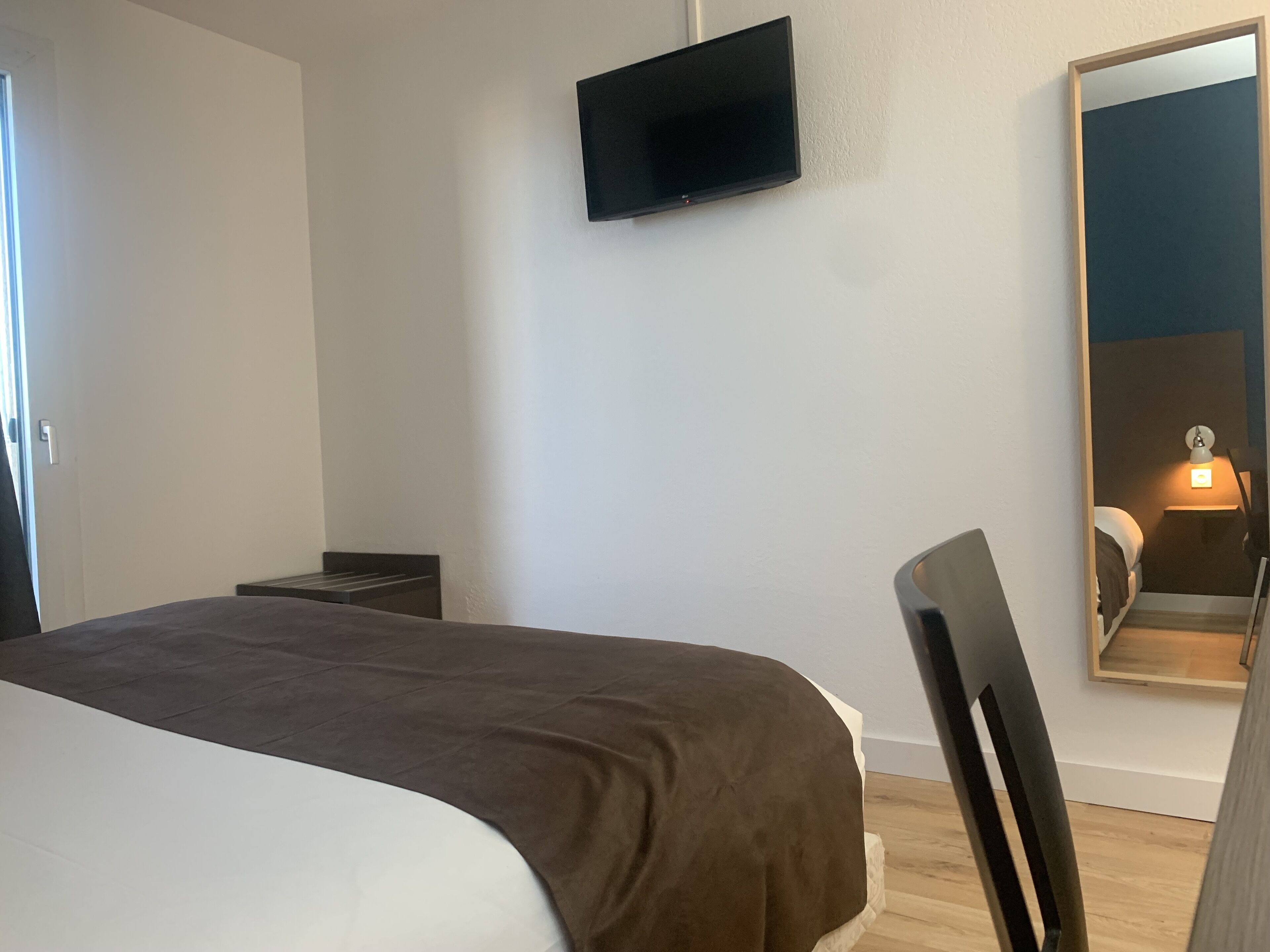 A hotel room with a bed, a TV mounted on the wall, a large mirror, and a bedside table with a lamp.