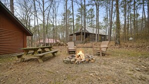 A rustic cabin with a fire pit in the center, surrounded by a wooden deck and a picnic table. - Crabapple Cove | Sleeps 8 | Porch Swing, Fire Pit & Near Beavers Bend (Broken Bow)