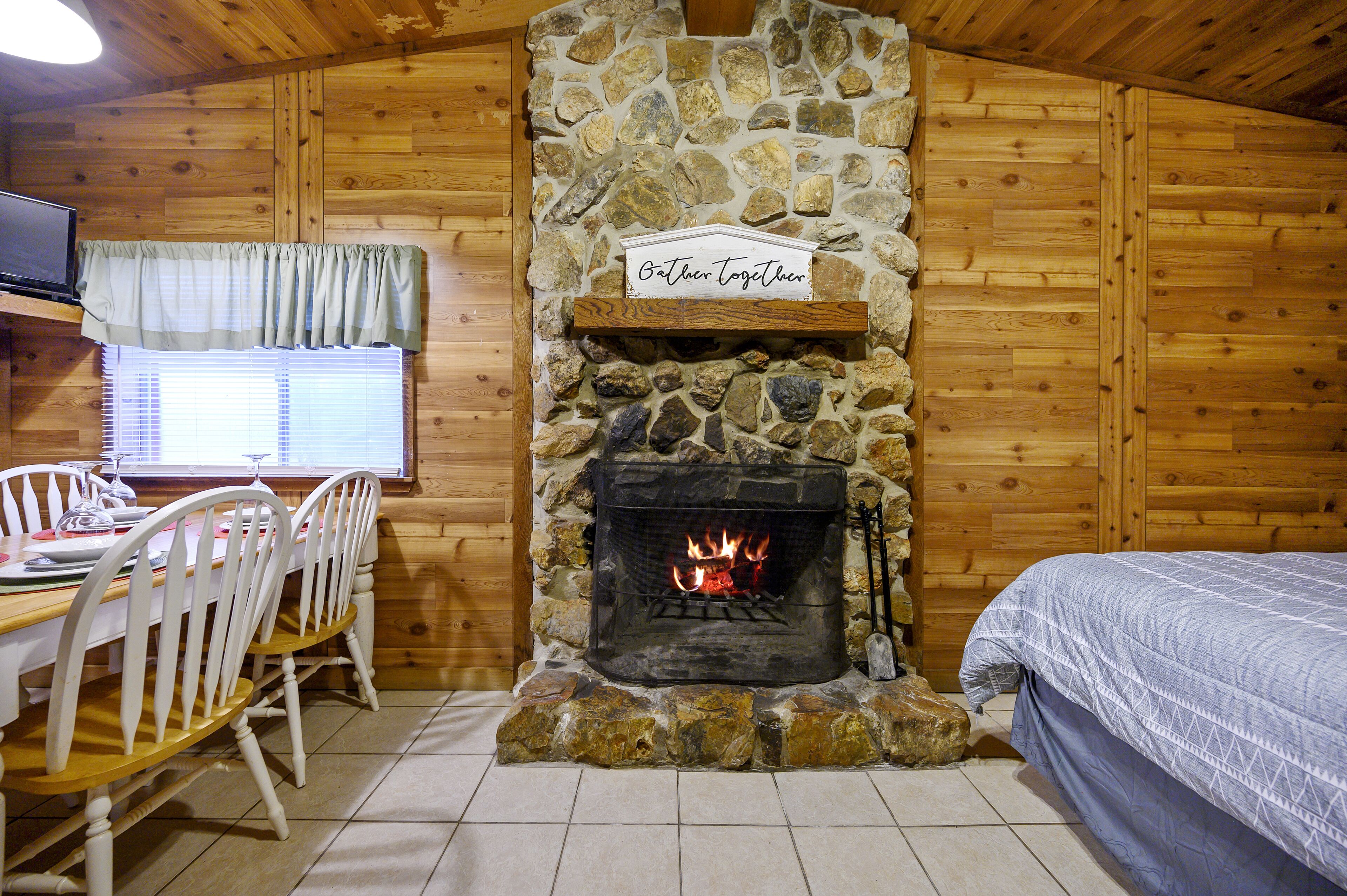 Une pièce chaleureuse avec une cheminée en pierre, des murs en bois, un coin repas avec des chaises et une table, et un lit recouvert d’une couverture à motifs.