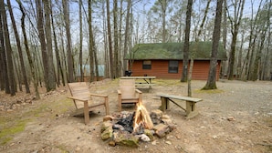 Una cabaña con un pozo de fuego rodeado de sillas de madera y una mesa de picnic, ubicada en un área boscosa.