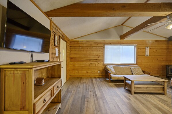 A cozy cabin interior with wooden walls, a television mounted on the wall, a wooden table, a sofa, and a window with blinds.