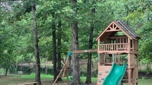 A wooden playground with a slide and climbing structure, surrounded by trees and a dirt ground.