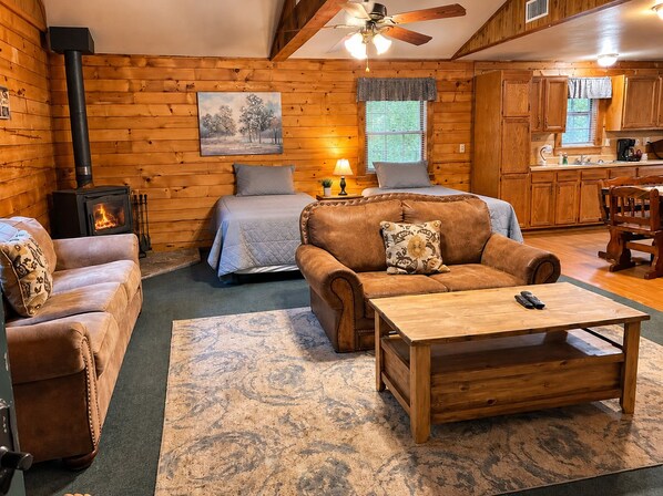 A cozy cabin interior with a bed, two sofas, a coffee table, a fireplace, and a kitchen area.