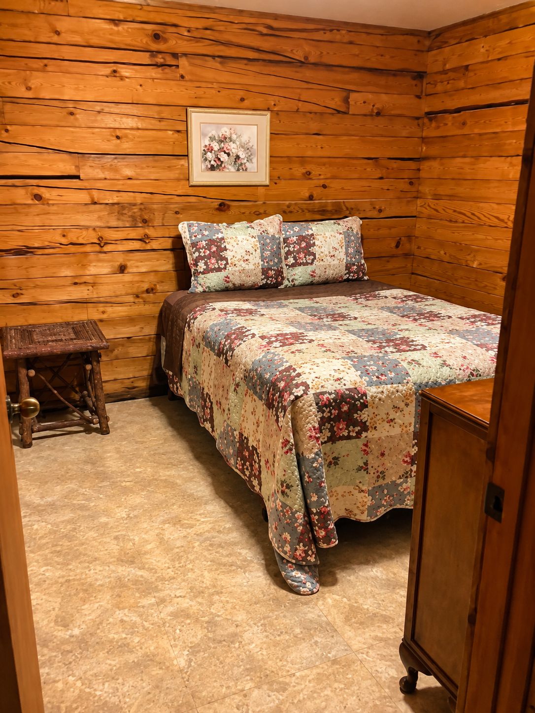 A bedroom with a wooden wall, a bed with floral bedding, a small wooden stool, and a wooden cabinet.
