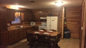A compact kitchen with wooden cabinets, a dining table set for four, a microwave, a refrigerator, and a trash bin.