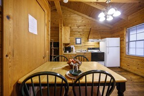 A wooden cabin interior with a dining table, chairs, a refrigerator, and a kitchen area. - Peach Blossom | Sleeps 6 | Pond View Deck, Fireplace & Near Beavers Bend (Broken Bow)