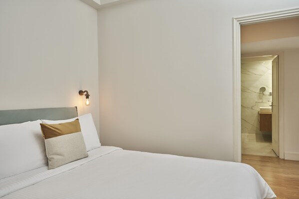 A neatly made bed with white and beige bedding, a headboard with a light-colored panel, and a wall-mounted light fixture.