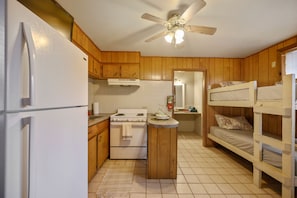 A compact kitchen with wooden cabinets, a refrigerator, a stove, and a small island. There are bunk beds in the adjacent room.