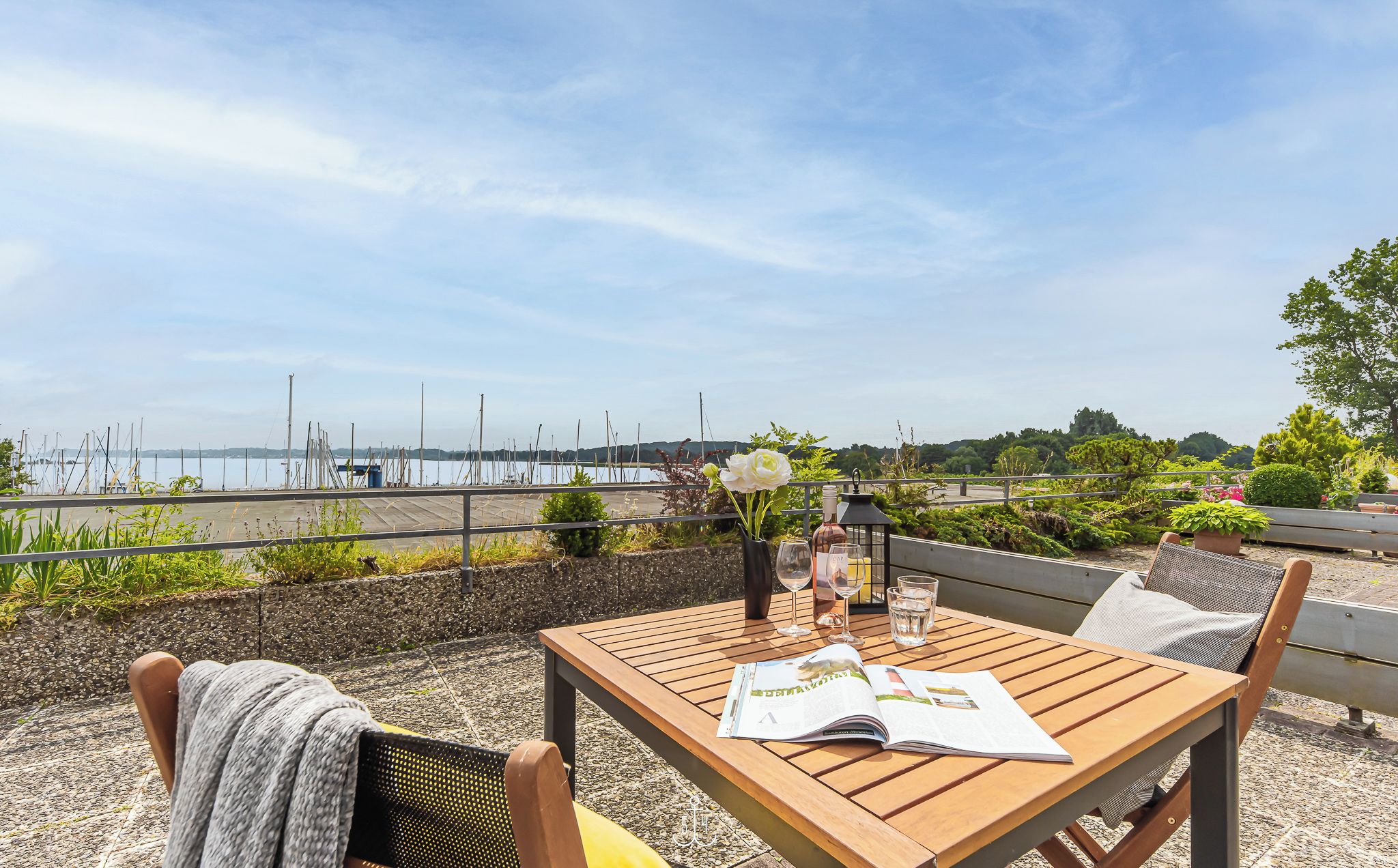 Ein Holztisch mit einer Zeitschrift, einer Blumenvase und einem Glas Wasser auf einer Terrasse mit Blick auf einen Yachthafen und Boote.