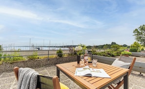 Ein Holztisch mit einer Zeitschrift, einer Blumenvase und einem Glas Wasser auf einer Terrasse mit Blick auf einen Yachthafen und Boote.