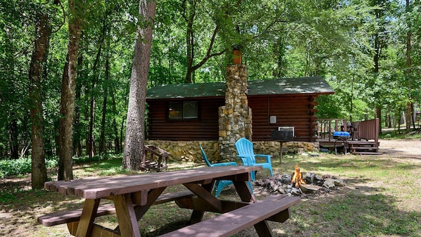 A rustic cabin with a stone chimney, surrounded by a forest. There is a picnic table with benches and a fire pit nearby. - Sweet Pea Place | Sleeps 4 | Fireplace, Fire Pit & Near Beavers Bend (Broken Bow)