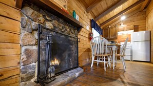 A rustic cabin interior with a stone fireplace, wooden dining table, and white chairs. - Sweet Pea Place | Sleeps 4 | Fireplace, Fire Pit & Near Beavers Bend (Broken Bow)