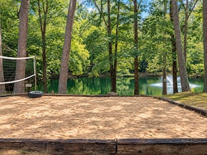 A sandy volleyball court with a net, surrounded by trees and a calm lake.