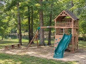 A playground with a wooden slide and swing set, surrounded by trees and a lake.