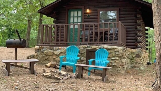 Una cabaña rústica con un porche, dos sillas Adirondack azules, una mesa y un estufa de leña.
