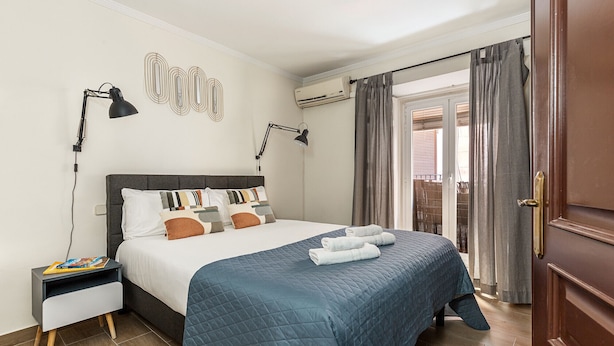 Une chambre d’hôtel avec un lit, des lampes de chevet, une table de nuit, un climatiseur fixé au mur et une vue sur l’extérieur, visible à travers une porte.