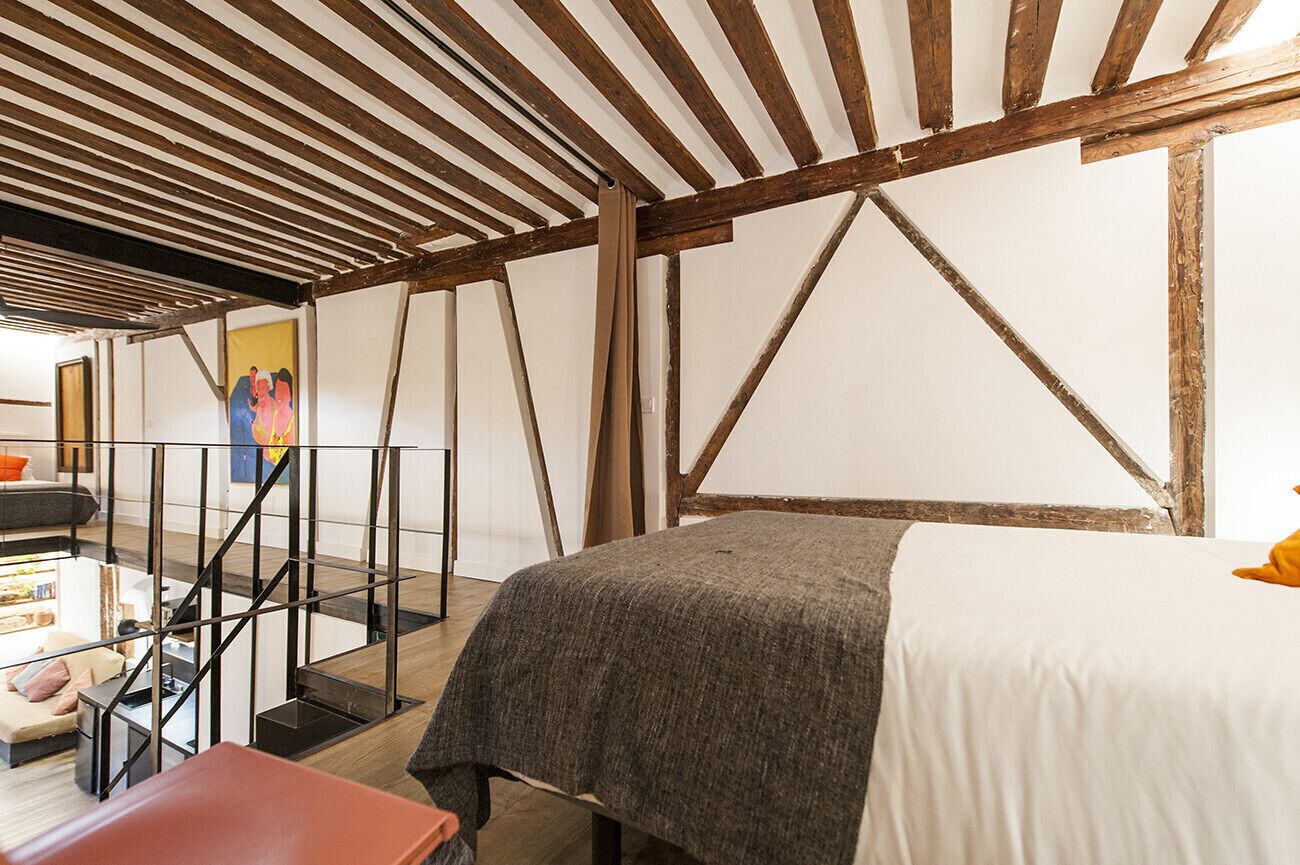 A room with exposed wooden beams, a bed with a white sheet, a sofa, and a staircase.