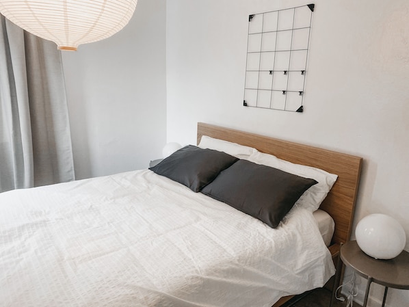 A neatly made bed with a white duvet and two dark pillows, a round white table with a white spherical object on it, and a wall-mounted light fixture.