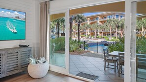 Habitación con televisión mostrando un barco en el agua, una planta en maceta, y un ventanal corredero que da a una zona de piscina con palmeras y un edificio al fondo.