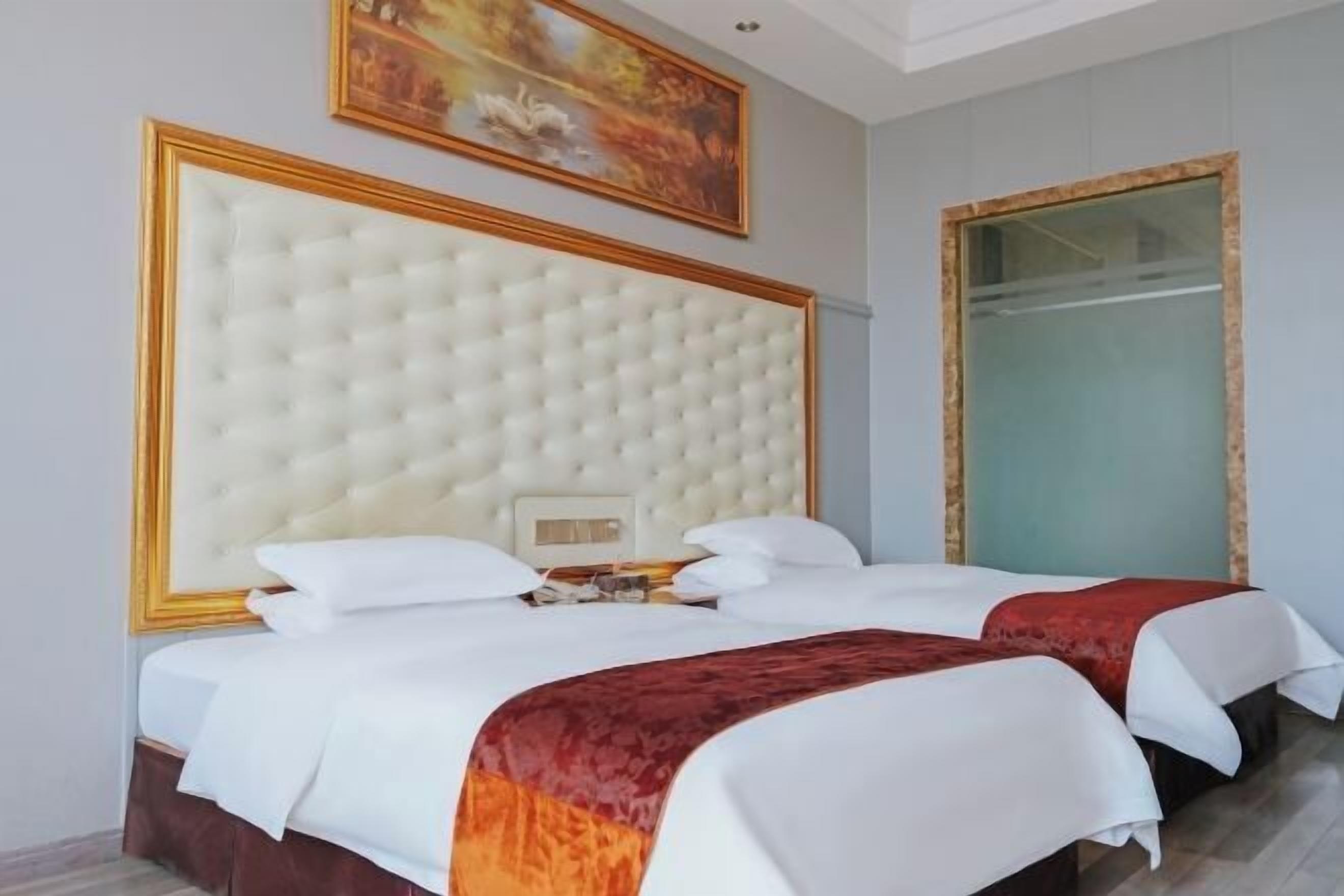 A hotel room with a large tufted headboard, two beds with white bedding, a red accent throw, a painting on the wall, and a large mirror.