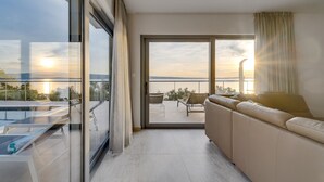 A modern living room with a sofa, a glass door leading to a balcony, and a view of the sunset over the water. - Seaview Villa Big Blue with 32sqm heated pool, 4 bedrooms (Podstrana)