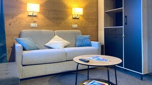 A modern room with a wooden wall, a grey sofa with cushions, a round wooden table with a remote control and magazines, and a wardrobe with open shelves.