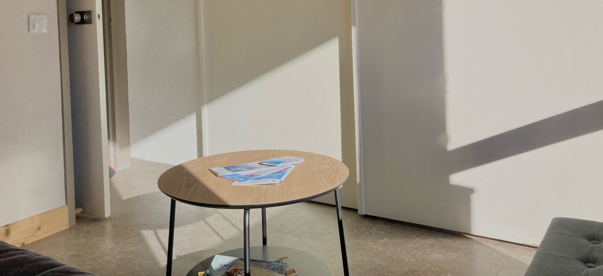 Una pequeña mesa redonda de madera con una superficie de cristal, ubicada en un rincón de una habitación con una puerta blanca y un suelo de tono claro.
