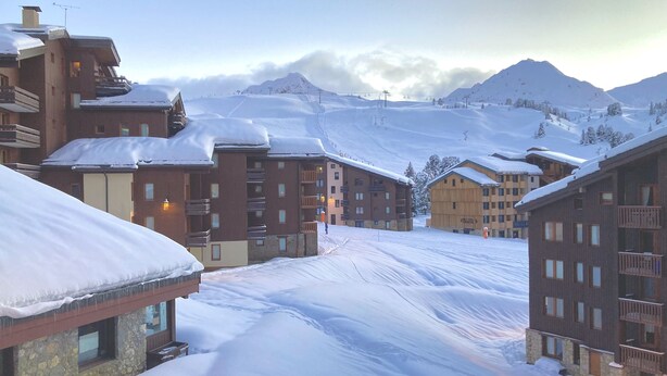 A snow-covered mountain resort with multi-story buildings, ski lifts, and a clear view of the mountains.