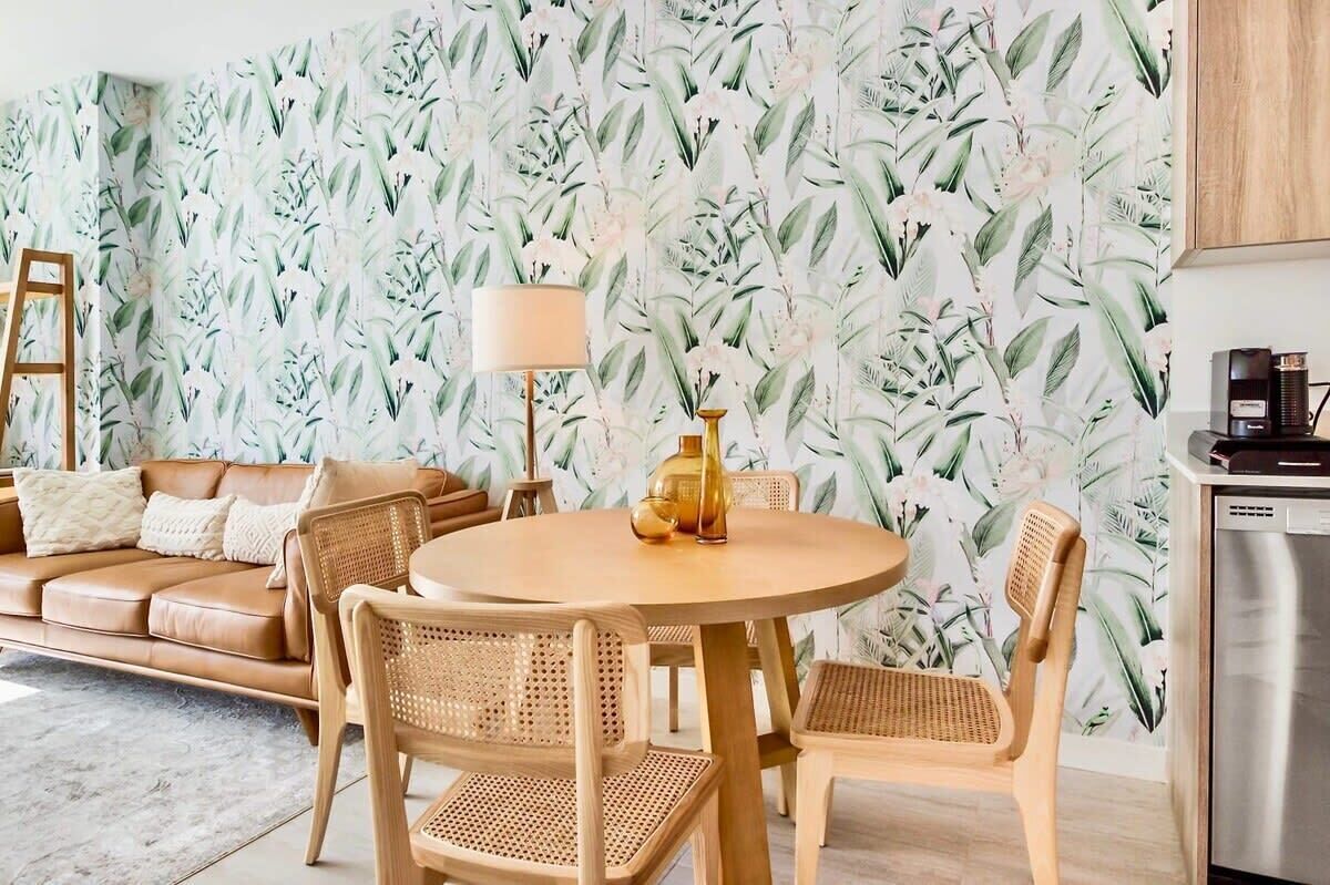 A living room with a round wooden table, wicker chairs, a brown leather sofa, and a wall with a tropical leaf pattern.