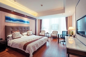 A hotel room with a large bed, a desk, two chairs, a TV, and a painting on the wall. - Henglong International Hotel (Karamay)