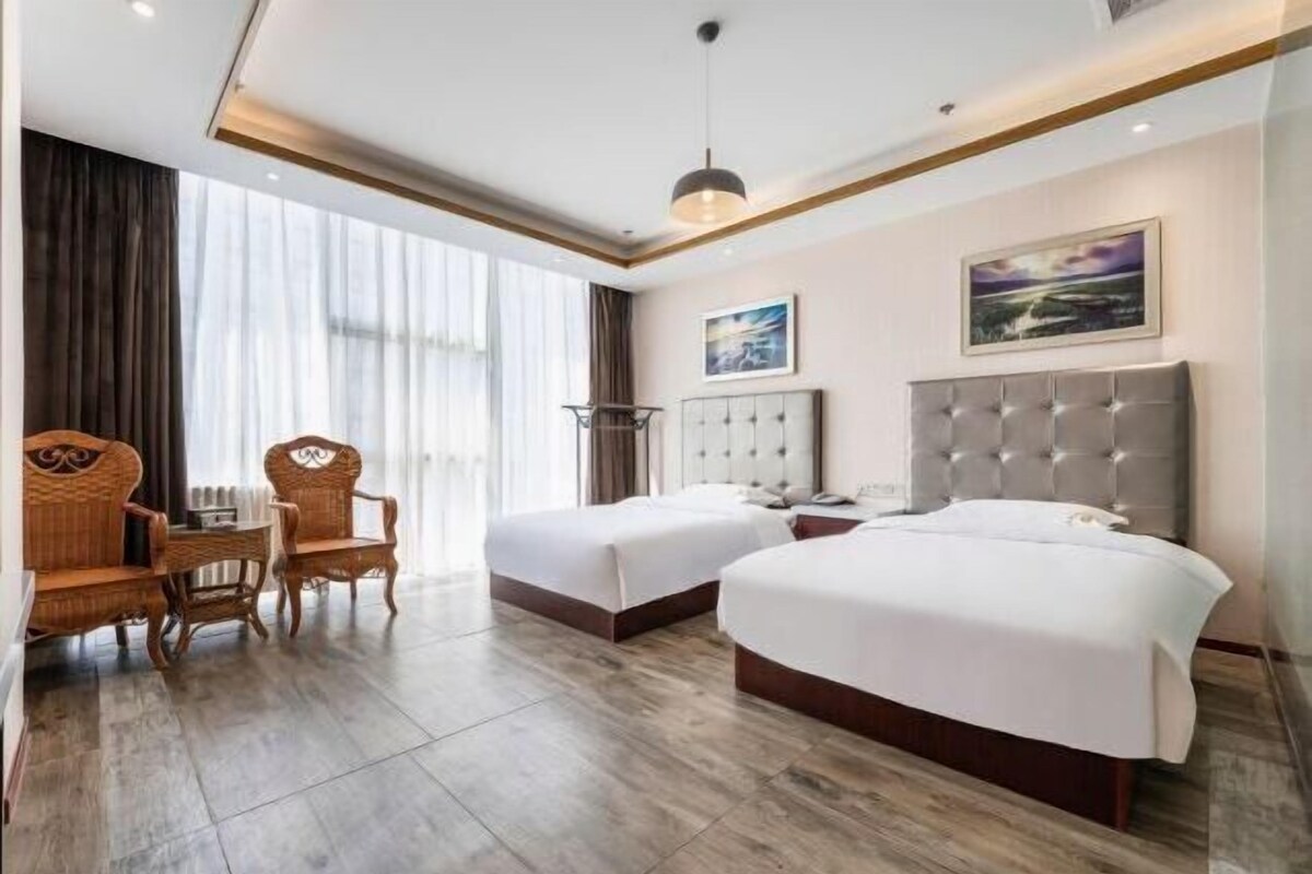Habitación de hotel con dos camas, una mesa de comedor con sillas, un ventanal con cortinas y un cuadro en la pared.