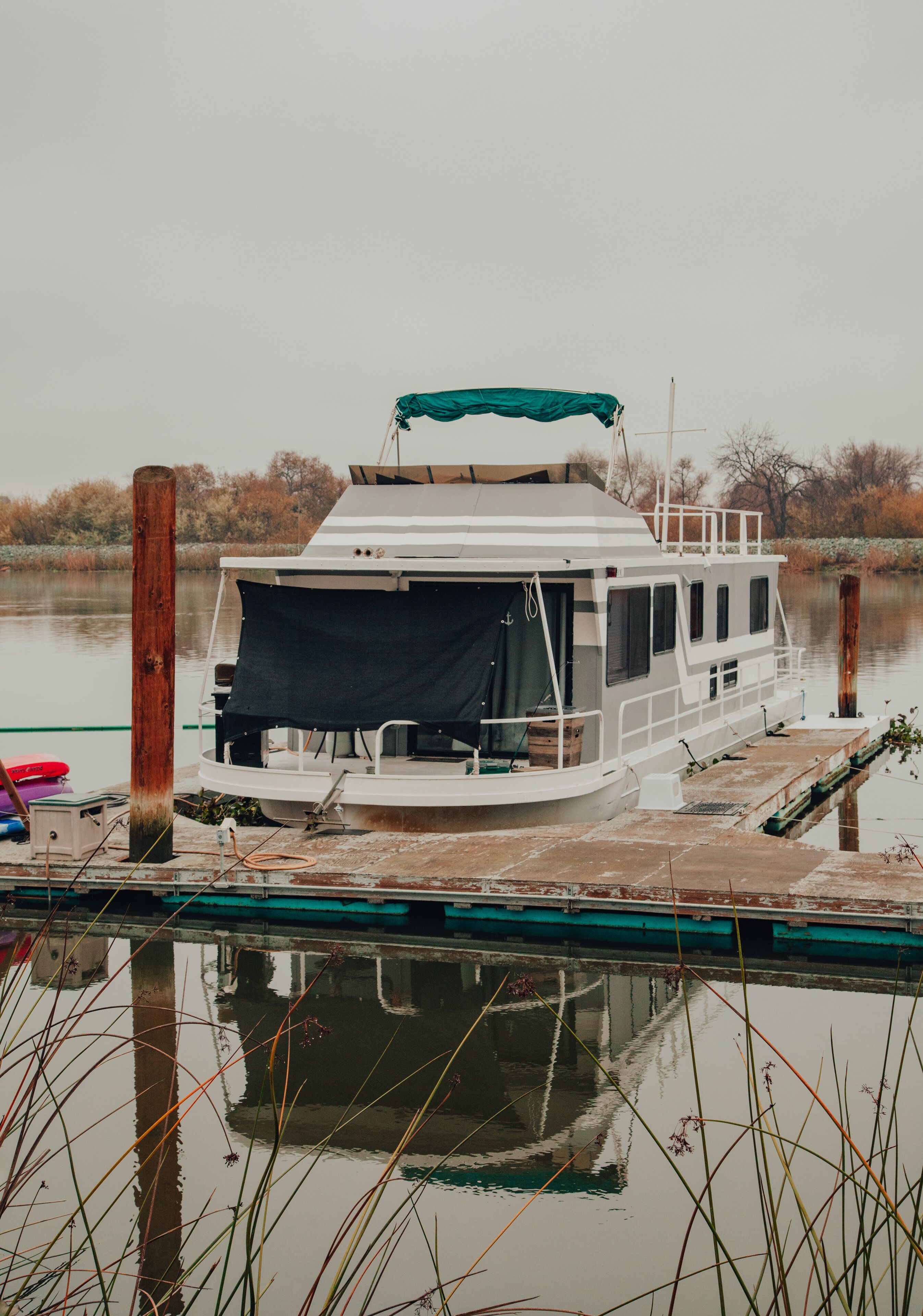 Ein Hausboot an einem Steg, mit einer überdachten Deckterrasse, Fenstern und einer kleinen Sitzgelegenheit im Freien.