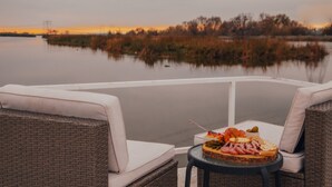 A dining table with a variety of food items, wicker chairs, and a view of a calm lake with trees in the background. - Romantic Getaway with Sauna, fireplace and great seal watching (Oakley)