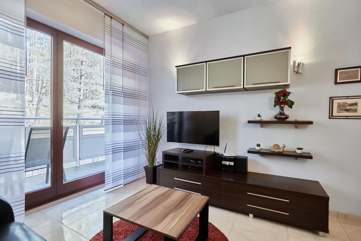 A modern living room with a large TV, dark wooden entertainment unit, and a glass door leading to a balcony.