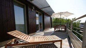 Eine Holzterrasse mit Liegestuhl und Tisch, eine Pergola mit Sonnenschirm und Blick auf ein Wohngebäude.